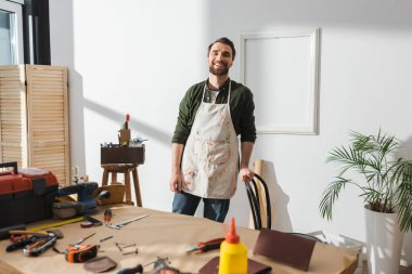 Smiling craftsman in apron standing near blurred tools on table 
