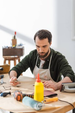 Craftsman in apron holding square tool near equipment in workshop 