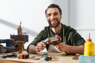 Cheerful carpenter holding electric screwdriver and looking at camera in workshop 