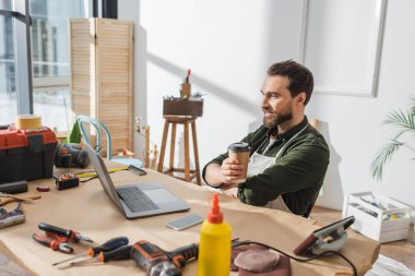 Smiling carpenter holding coffee to go near devices and tools in workshop  clipart