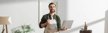 Cheerful carpenter in apron holding laptop and looking at camera in workshop, banner 