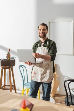 Happy craftsman in apron holding laptop near chairs in workshop 
