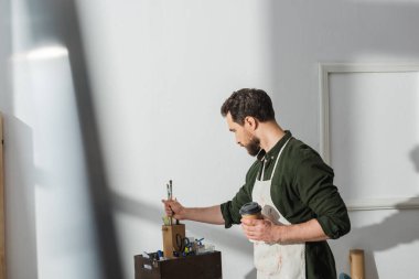 Side view of carpenter holding coffee to go and taking paintbrushes in workshop 