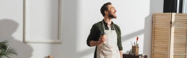 Smiling bearded repairman in apron holding paper cup in workshop, banner 