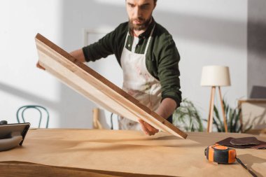 Blurred carpenter holding wooden board near table in workshop 