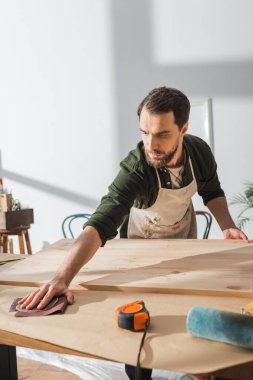 Bearded carpenter taking sandpaper while working with wooden board in workshop  clipart