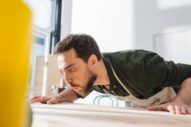 Bearded carpenter in apron blowing away dust on wooden board