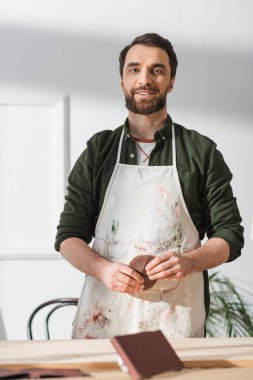 Smiling carpenter holding sandpaper near blurred wooden board in workshop 