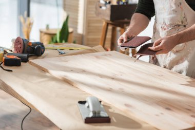 Cropped view of craftsman holding sandpaper near wooden board and tools