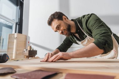 Smiling bearded workman sanding wooden board in workshop 