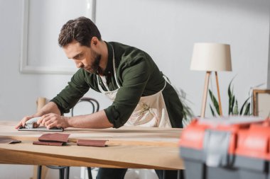 Repairman in apron sanding wooden plank near blurred toolbox in workshop  clipart