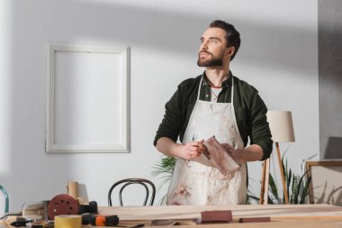 Bearded craftsman in apron holding sandpaper near blurred wooden board on table 