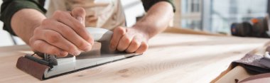 Cropped view of man in blurred apron sanding wooden board in workshop, banner  clipart