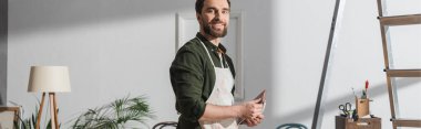 Cheerful restorer in apron holding sandpaper and looking at camera in workshop, banner 