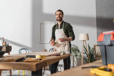 Smiling restorer in apron holding ruler and looking at camera near wooden board in workshop 