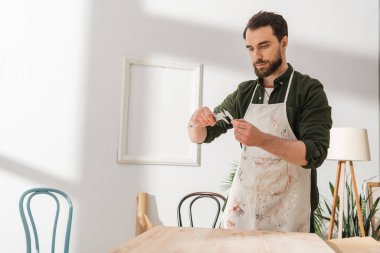Bearded carpenter in apron using calipers while working with wooden board in workshop 