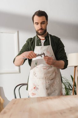 Bearded restorer in apron looking at calipers near blurred wooden board 