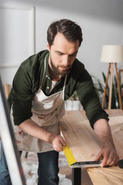 Bearded carpenter using square tool on wooden board in workshop 