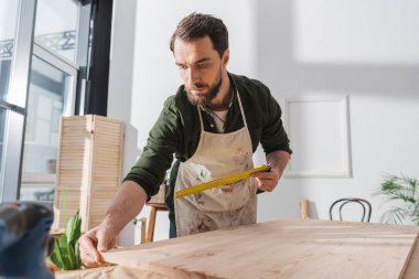 Carpenter in apron holding square tool and touching wooden board in workshop 