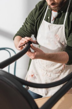 Cropped view of restorer in apron sanding part of chair in workshop 