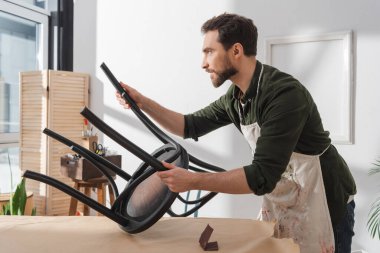 Side view of restorer in apron holding chair near sandpaper on table in workshop 