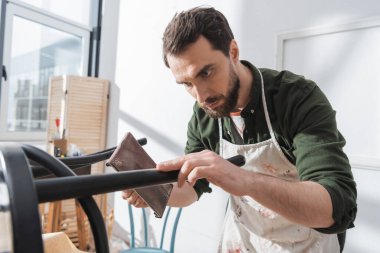 Restorer in dirty apron sanding paint on wooden chair in workshop 