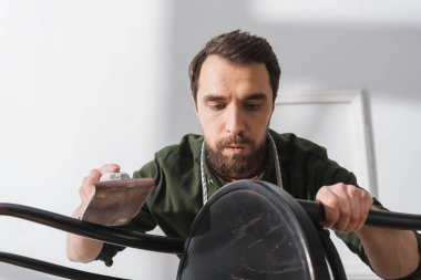 Restorer holding sandpaper and blowing on wooden chair in workshop 