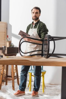 Restorer holding sandpaper and looking at camera near wooden chair in workshop 
