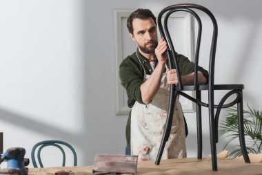 Bearded craftsman in apron looking at wooden chair near sandpaper and sanding machine in workshop 