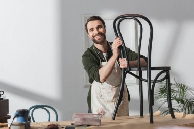 Cheerful restorer in apron looking at camera near chair and sandpaper in workshop 