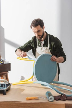 Craftsman in apron repainting chair near roller on table in workshop  clipart