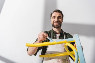 Smiling bearded restorer looking at camera while repainting chair in workshop 