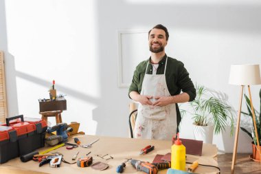 Positive restorer in apron looking at camera near tools on table in workshop 
