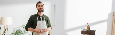 Smiling restorer in apron looking at camera in workshop, banner 