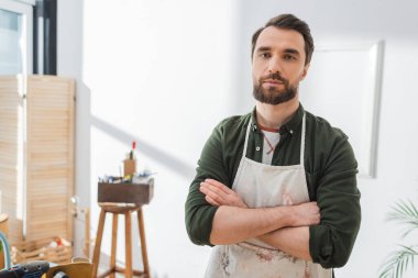 Bearded craftsman in apron crossing arms in workshop 