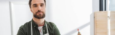 Bearded restorer looking at camera while standing in workshop, banner 