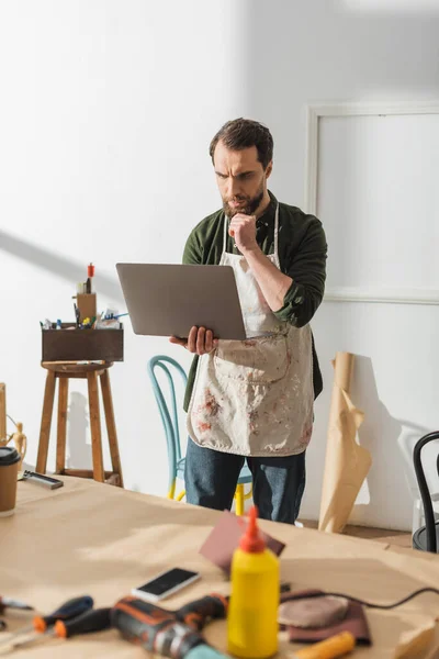 stock image Pensive carpenter in apron using laptop in workshop 