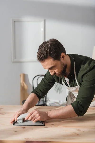 stock image Bearded carpenter in apron sanding wooden board 