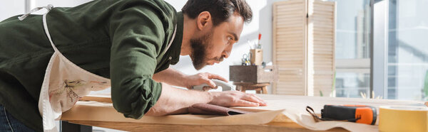 Side view of bearded carpenter blowing dust from wooden board in workshop, banner 