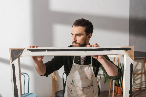 stock image Bearded carpenter in apron looking at picture frame in workshop 