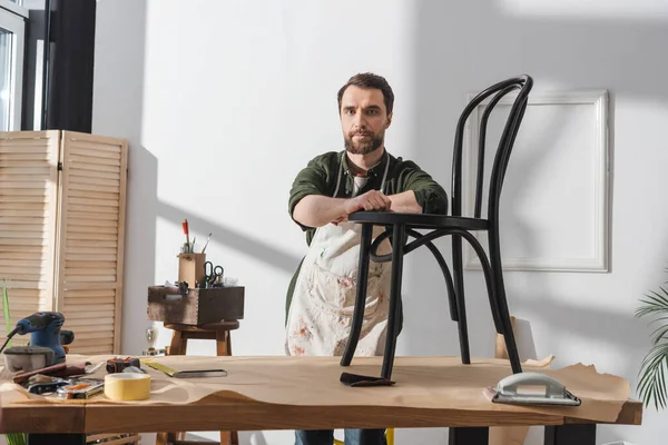 stock image Bearded restorer in apron looking at camera near wooden chair in workshop 