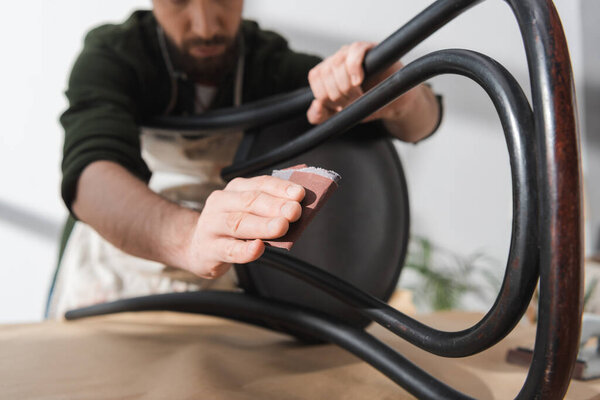 Cropped view of blurred repairman rubbing chair with sandpaper in workshop 