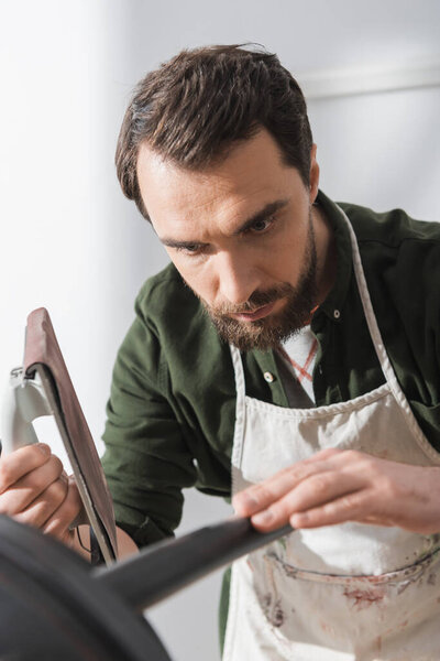 Focused restorer holding sandpaper near blurred chair in workshop 