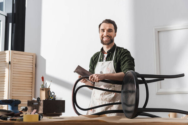 Smiling restorer in apron holding sandpaper near chair and looking at camera in workshop 