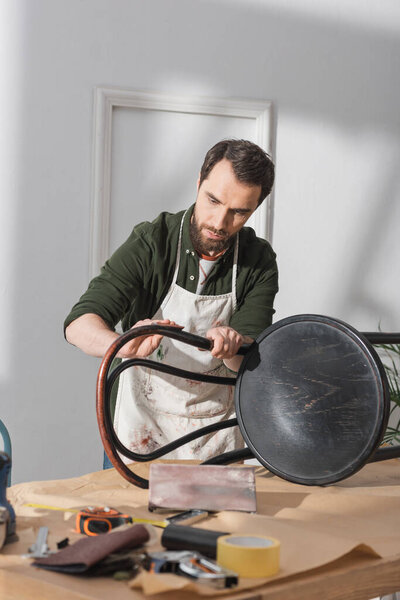 Bearded restorer sanding chair near blurred tools on table in workshop 
