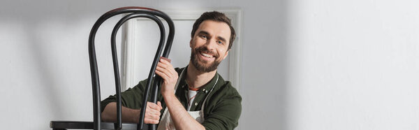 Positive restorer in apron holding chair and looking at camera in workshop, banner 