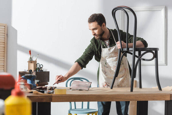 Bearded restorer in apron holding taking sandpaper near chair on table in workshop 