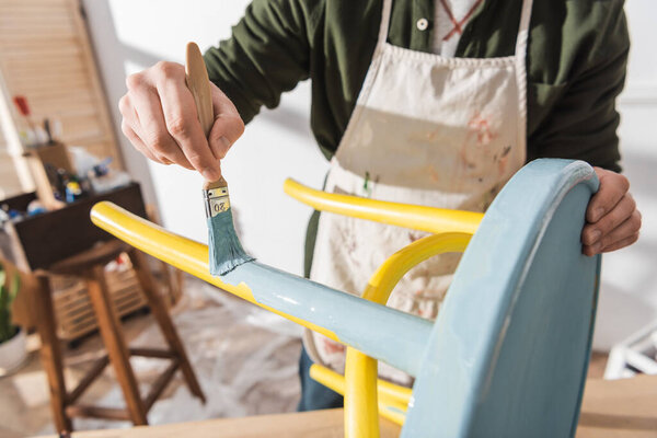 Cropped view of craftsman painting on wooden chair in blurred workshop 