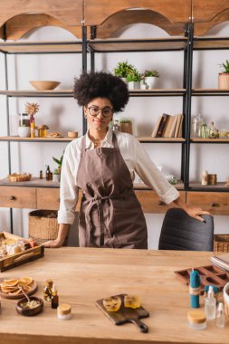 smiling african american woman in eyeglasses and apron standing near natural ingredients for soap and candle making