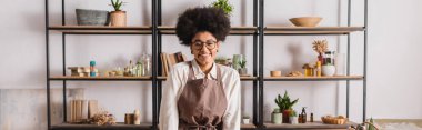 happy african american woman in eyeglasses and apron standing near blurred rack with books and natural ingredients in craft workshop, banner